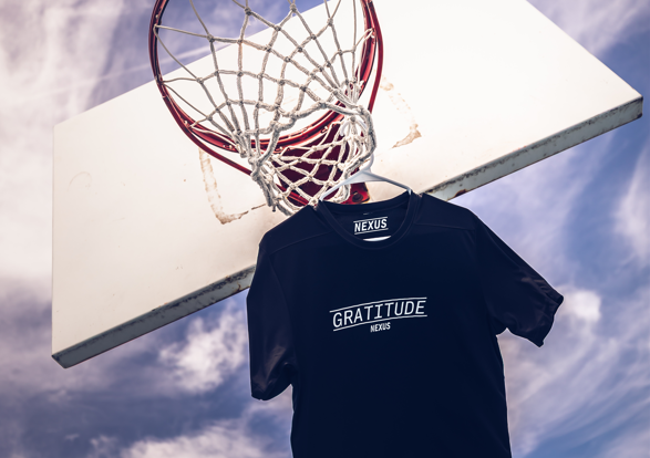 Black t-shirt with 'Gratitude' text hanging on a basketball hoop against a cloudy sky.